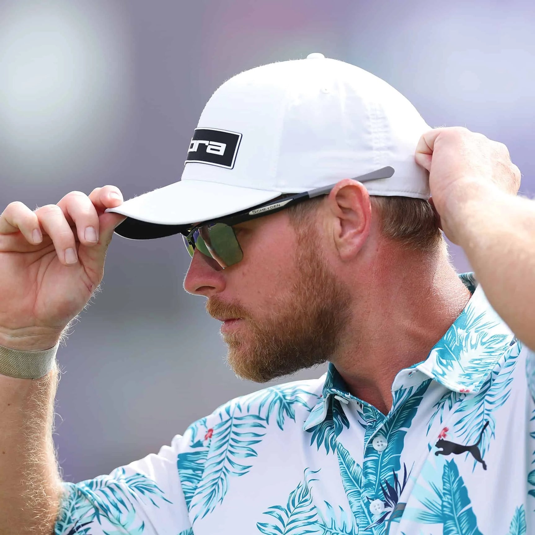 A man in a patterned palm leaf shirt adjusts his white cap while wearing Scheyden CIA GRABBER Sunglasses, size 65.