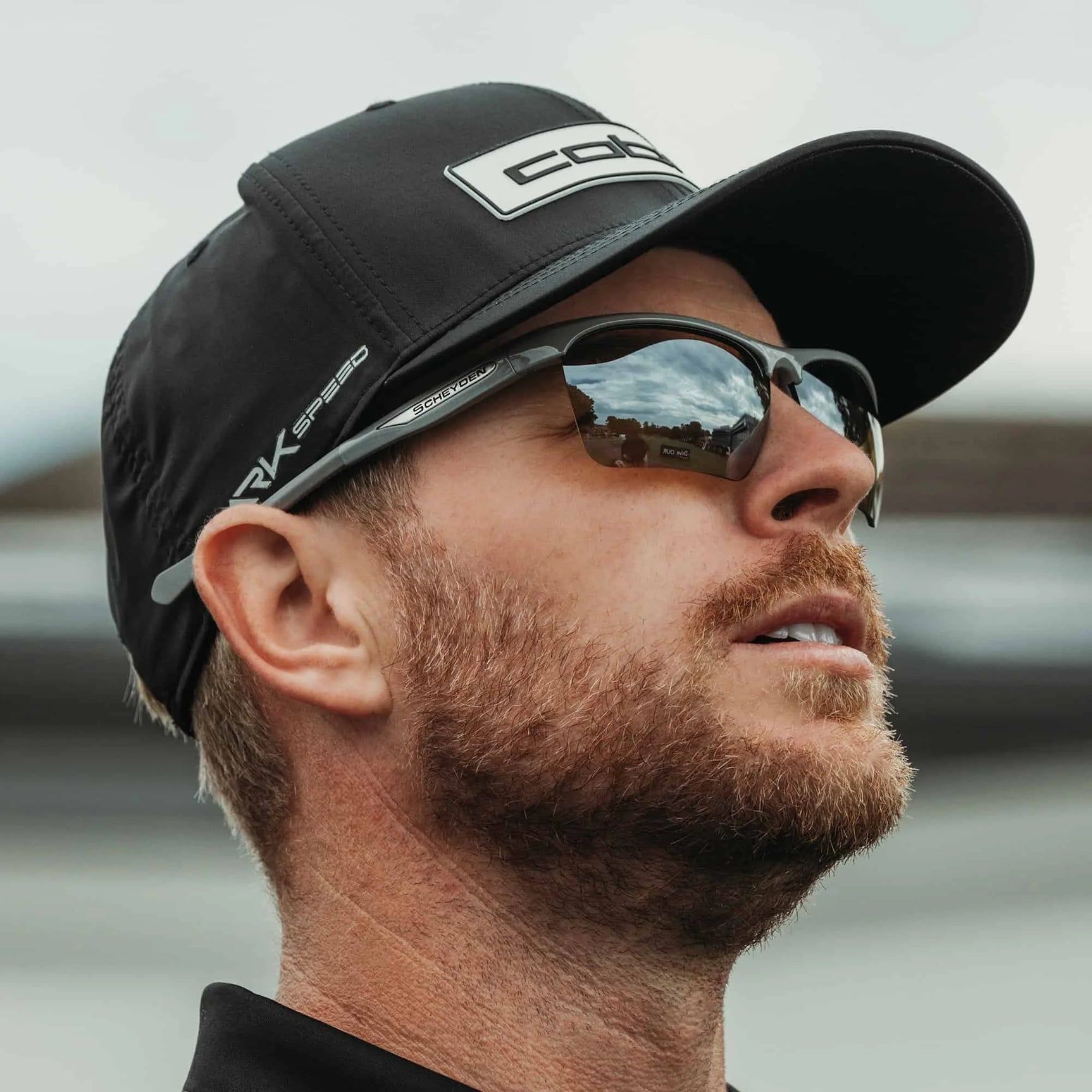 A bearded man, wearing a cap, gazes forward with the sky mirrored in his Scheyden CIA GRABBER Sunglasses. A bearded man, wearing a cap, gazes forward with the sky mirrored in his Scheyden CIA GRABBER Sunglasses.
