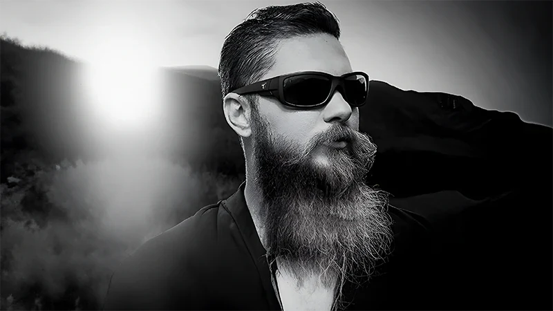 Man with a long beard and sunglasses outdoors, with sunlight shining in the background, in black and white.
